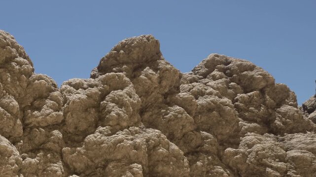 Large rock formations rise against a clear blue sky in a desert scene. The texture of the rocks is visible, showing natural shapes and structures of the landscape.