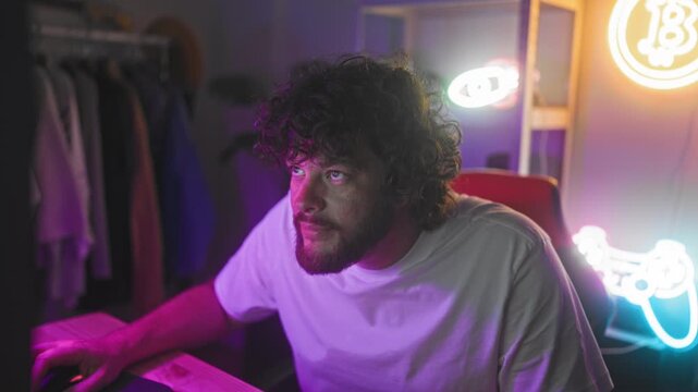 Man with beard and curly hair raises fist while gripping controller in gaming studio with neon sign and desk setup; triumph.