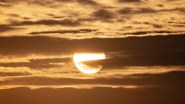Timelapse of a big glowing sun rising through dramatic golden clouds at sunset or sunrise. Beautiful warm cinematic footage fits morning news intros or corporate video headers as a design element.