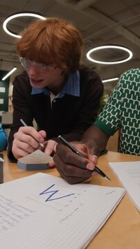 Vertical video of Caucasian young adult male with glasses collaborating with African American colleague writing in notebook. Clip is ideal for corporate training or university recruitment ads