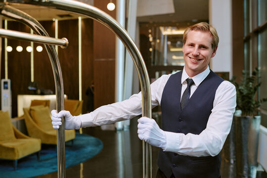 Portrait of smiling young adult Caucasian bellhop holding luggage cart in hotel lobby. Image represents hospitality industry and is suitable for luxury tourism marketing