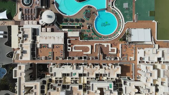 White resort buildings form geometric rooftop patterns around courtyards and terraces, aerial top-down overhead tracking over pools and architecture