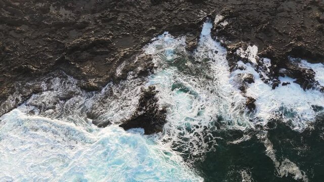 White ocean surf crashes over jagged dark volcanic rocks along a rugged coast, aerial overhead zenithal static background with whitewash and mist spray