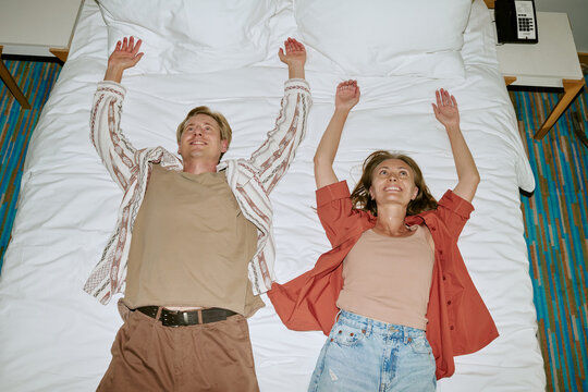 Happy Caucasian young adult couple lying on bed in hotel room with hands up, smiling. High angle view of cheerful tourists relaxing during vacation, perfect for travel agency marketing