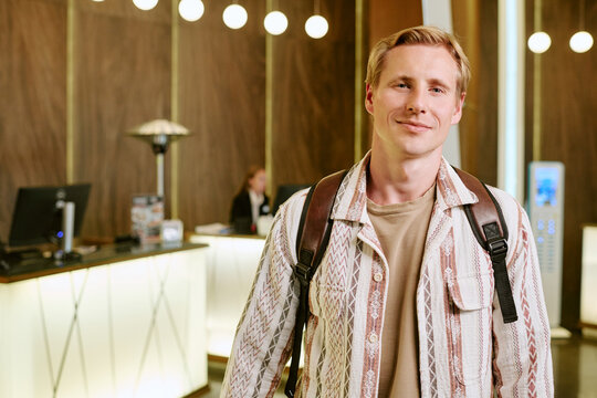 Portrait of smiling young adult Caucasian man with backpack standing in hotel lobby. Image represents travel, hospitality, and tourism, suitable for booking platform advertisements