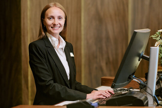 Portrait of smiling young adult Caucasian woman working at hotel reception desk using computer. Image represents hospitality industry and is suitable for customer service marketing