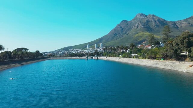 Drone shot over a large blue reservoir lined with palm trees, with Cape Town&rsquo;s city apartment towers and suburban homes visible along the shoreline and the rugged slopes of Devil&rsquo;s Peak rising behind.