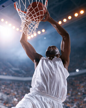 Athlete leaps toward basket with passion. Crowded arena pulses with electric energy. Light beams cut through hazy, atmospheric air. Focus, strength, and grace define his motion