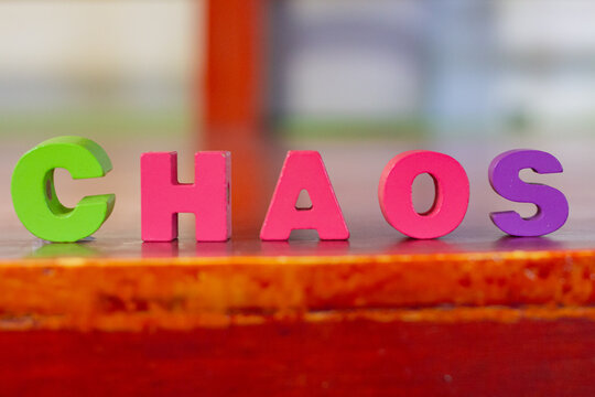Colorful wooden letter blocks spelling the word CHAOS arranged in a row on a textured wooden surface against a blurry background.