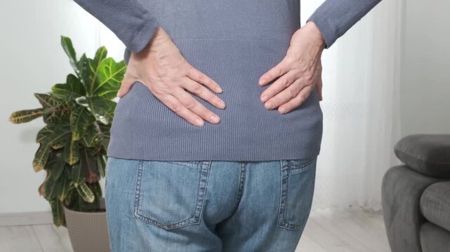 Back pain. Middle aged woman holding her lower back and massaging it with her hands at home. Close-up, rear view, side view. Lumbago, sciatica, lower back pain, lumbago in the back, hernia, protrusion