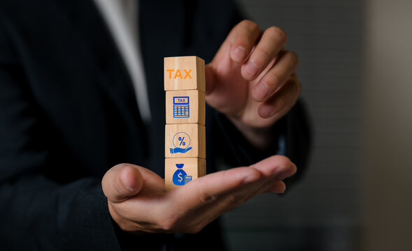 A close-up of a person in formal attire holding wooden blocks with icons representing tax, percentage, and disability, illustrating financial concepts and business planning.