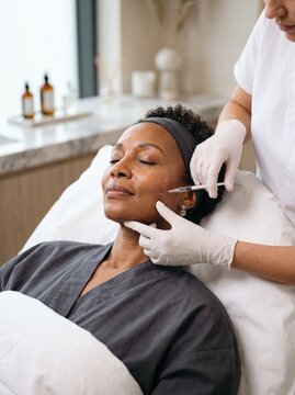 A senior woman with gray hair receives a dermal filler injection near her jaw from a gloved specialist, a cosmetic treatment concept.