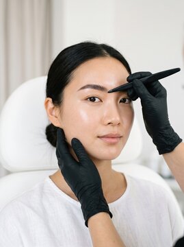 An Asian woman has her eyebrows shaped with tweezers by a gloved specialist, a beauty salon eyebrow treatment concept.