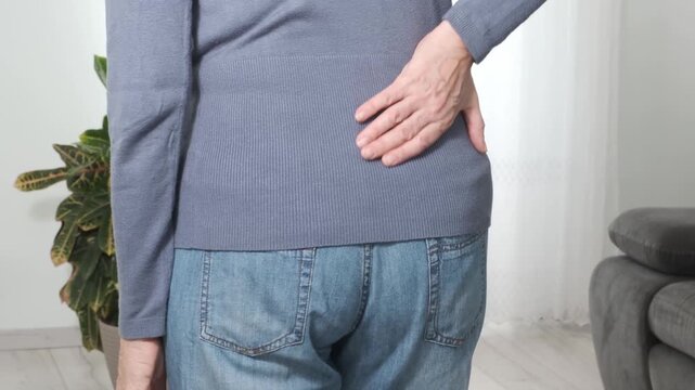 Back pain. Middle aged woman holding her lower back and massaging it with her hands at home. Close-up, rear view, side view. Lumbago, sciatica, lower back pain, lumbago in the back, hernia, protrusion