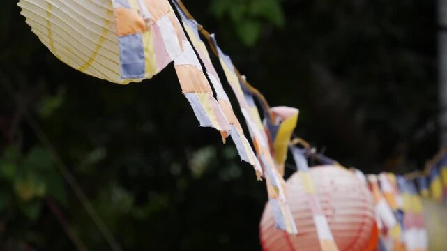 Colorful buddhist prayer flags and hanging paper lanterns waving in the wind against a green blur. Sacred symbols.