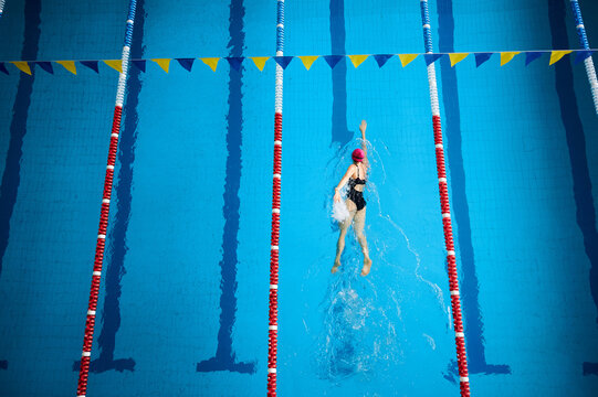 Professional swimmer training in pool lane, performing freestyle stroke. View from above. Athlete in pink cap and black swimsuit, blue water with lane dividers, sport and endurance concept