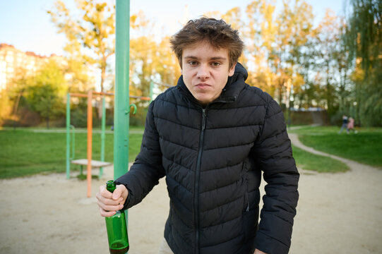 Teen boy with bottle poses in public recreation space. Harsh look and uneasy mood create realistic scene of pressure and risky behavior. Concept of underage alcohol abuse among minors