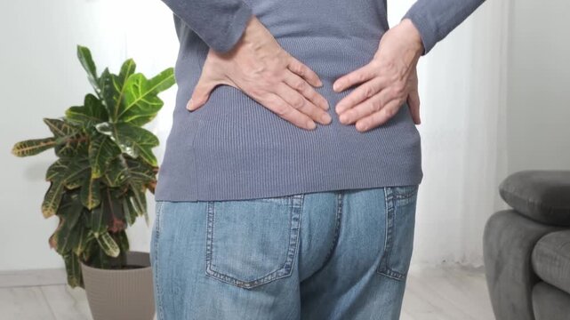 Back pain. Middle aged woman holding her lower back and massaging it with her hands at home. Close-up, rear view, side view. Lumbago, sciatica, lower back pain, lumbago in the back, hernia, protrusion