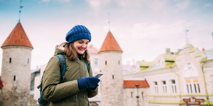 Tallinn, Estonia. Young Caucasian Woman Backpacker Tourist Enjoying Life, Smiling And Using Smartphone Near Landmark Viru Gate. Vacation In Old Town. Girl Surfing Net Internet In Phone