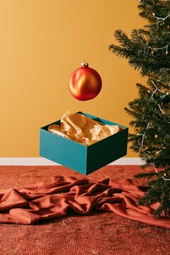 Glass Christmas ornament floating above an open blue gift box, glowing with warm amber light on a cozy rug for holiday retail and advertising copy space