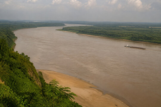 View from high bluff of wide brown river bending through low green forest, sandbar, tugboat and barge downstream, Mississippi River landscape