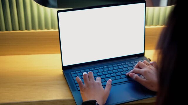 Hands type on laptop with blank screen. Warm light illuminates wooden desk and keyboard. Person focuses intently on work or study. Cozy setting suggests comfort and concentration