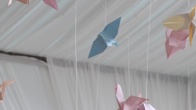 Origami cranes gently sway in slow motion, multicolored paper birds hanging from ceiling on strings, creating peaceful and decorative footage.