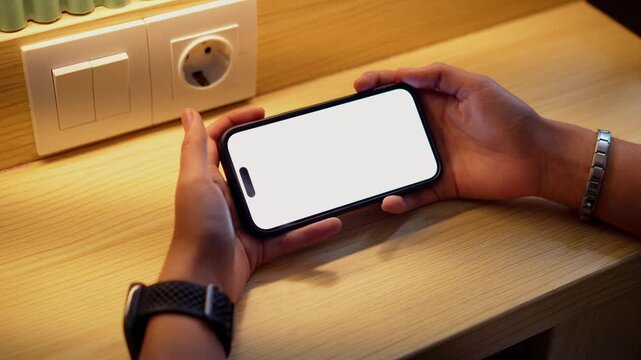 Hand holds phone with white screen. Phone rests on wooden surface near wall outlet. Warm light illuminates the scene. User taps screen with finger. Natural lighting adds cozy atmosphere