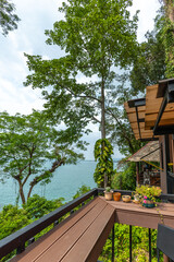 Fototapeta premium Wooden balcony terrace with bench and potted plants overlooking tropical sea and green forest. Coastal resort view, vacation destination, outdoor architecture, travel background, nobody.