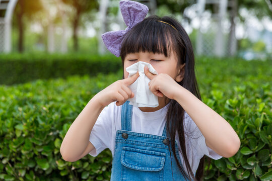 Asian little girl blowing nose with tissue outdoors, child with cold or allergy symptoms in park, healthcare and hygiene concept