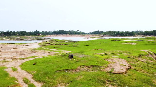 Two friend sit on a grassy pastoral field at riverbank with patches of bare soil, overlooking a wide river and distant tree line under a clear sky in an aerial view.
