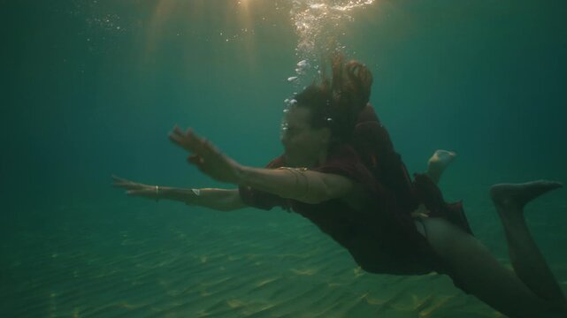 A woman swims underwater, her flowing dress trailing as sunbeams filter through the water.