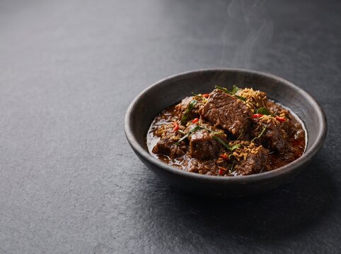 Traditional Indonesian beef rendang in dark ceramic bowl with lime leaves and toasted coconut, steaming on dark stone