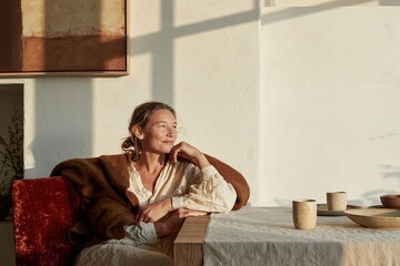 Fototapeta premium Mature woman resting in a cozy dining room, wearing a warm wool blanket with a thoughtful expression, featuring soft light and copy space