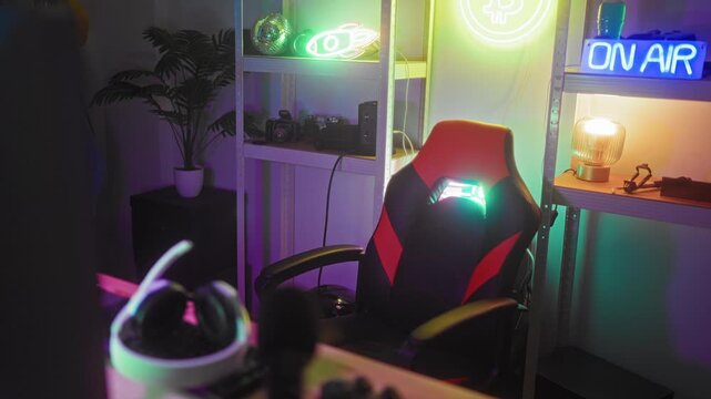 Man adjusts white headset and types on keyboard at a computer desk in studio with neon on air sign, gaming chair and shelf lighting; concentration.