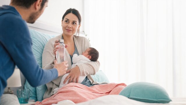 Man giving water bottle to woman holding newborn baby in bed at home. Postpartum care and hydration support for mother during recovery.