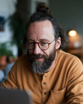 Man with glasses and beard wears warm-toned shirt. He looks focused, perhaps working or thinking deeply. Soft lighting creates a cozy, intimate atmosphere