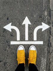 Fototapeta premium Person in Yellow Sneakers Standing Before Three Directional Arrows on Asphalt Representing Choice and Decision Making