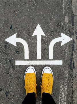 Person in Yellow Sneakers Standing Before Three Directional Arrows on Asphalt Representing Choice and Decision Making