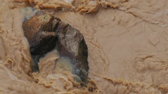 Natural hazards during the rainy season river surges and mudslides rapidly flowing muddy brown water rushes past dark scattered rocks weeds and trees within the stream or river.