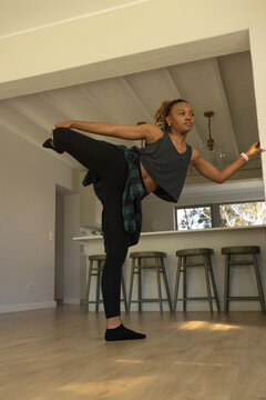 Woman balancing on one leg, holding foot, wearing crop top and watch beside kitchen island