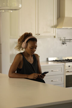 African American woman sitting at kitchen island using smartphone while wearing pink headphones