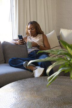 African American female sitting on sofa holding phone and green mug wearing sleeveless top at home