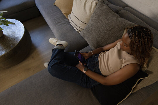 Adult African American woman reclining on gray sofa in living room holding phone