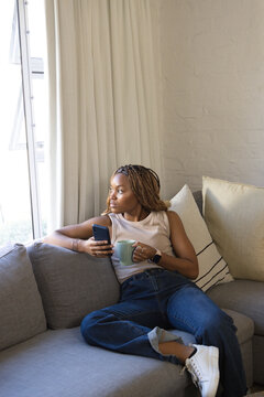 African American woman sitting on sofa at home in sleeveless top, jeans, holding smartphone and mug