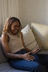 African woman sitting on light-colored sofa holding tablet and smiling at screen wearing beige tank © WavebreakMediaMicro