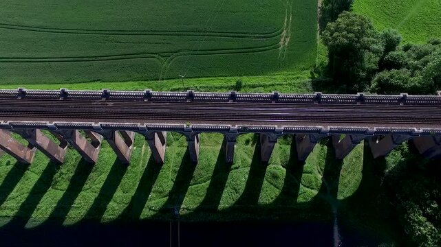 Drone ariel top-down view of a historic brick railway viaduct crossing lush green countryside, showcasing its repeating arches, railway tracks, fields and surrounding rural landscape