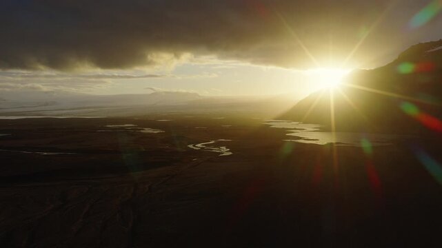 Volcanic wetlands at Herdisarvik, Olfus, South Iceland, extend toward the Atlantic Ocean with winding channels, dark lava terrain and golden sunlight through heavy clouds, drone forward flyover.