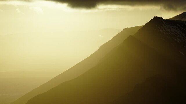 Volcanic mountain ridge rises above hazy coastal terrain in Iceland, with steep dark slopes illuminated by low golden sunlight and distant landforms fading into atmospheric haze, aerial view.