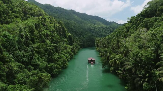 Drone ascends while following a Loboc River cruise boat downstream through green water and forested hills in Bohol